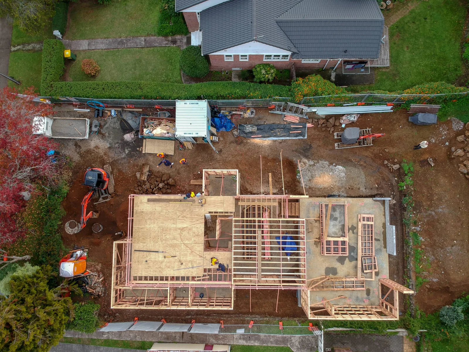 Aerial view of a construction site with a house frame being built, surrounded by green lawns and other houses
